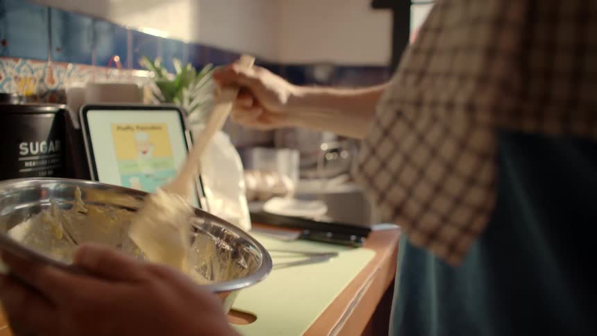 A young man happily follows a recipe on his tablet while mixing batter in a sunlit kitchen