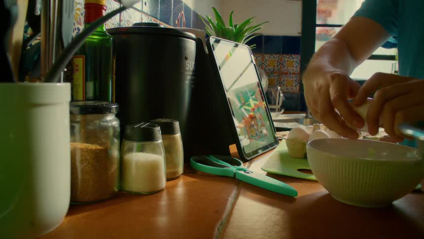 A person follows a recipe on a tablet while cracking an egg into a bowl in a sunlit kitchen