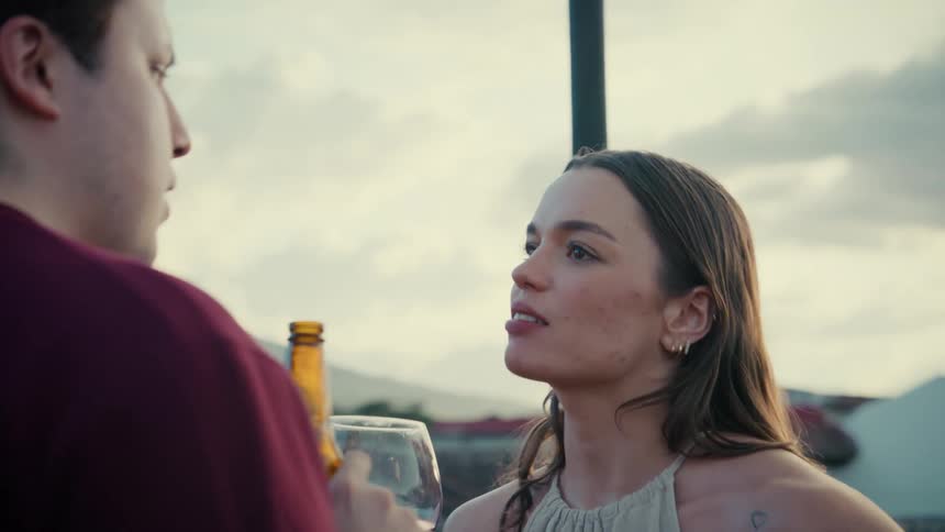 A young woman holds a glass of red wine while listening intently to a man who is drinking a beer during a conversation on a rooftop at dusk