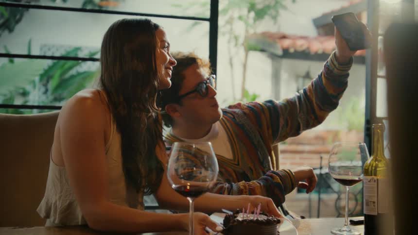 A happy young couple takes playful selfies together as they celebrate a birthday with a chocolate cake and glasses of red wine
