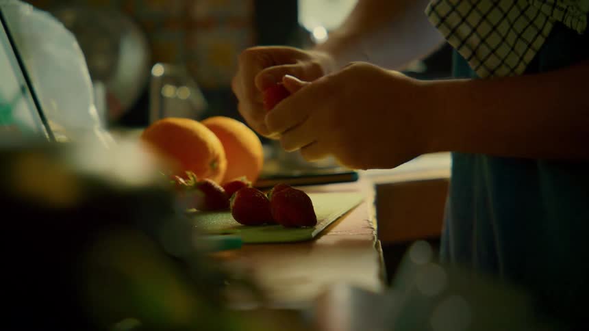 Close-up of a person's hands hulling fresh strawberries on a cutting board in a warmly lit kitchen