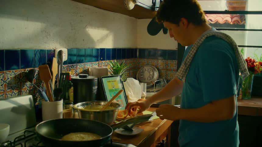 A young man follows a recipe on a tablet while cooking pancakes and preparing fresh fruit in a sunlit, rustic kitchen