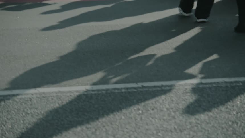 A low-angle slow-motion shot captures the feet and shadows of a crowd of anonymous pedestrians crossing a busy city street