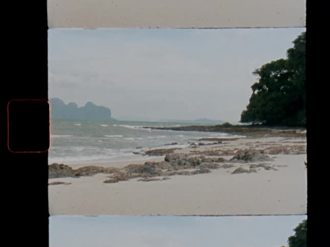 A vintage Super 8 film scan captures the waves washing up on a rocky tropical beach on an overcast day