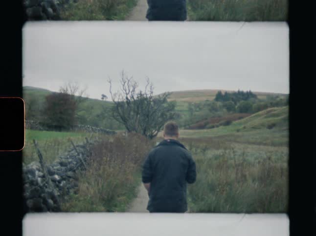 Vintage 8mm film captures a man from behind as he walks down a path through the rolling hills of the countryside