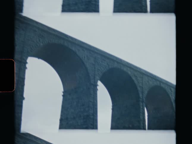 An archival Super 8 film captures the imposing architecture of a multi-arched stone bridge on a gloomy day