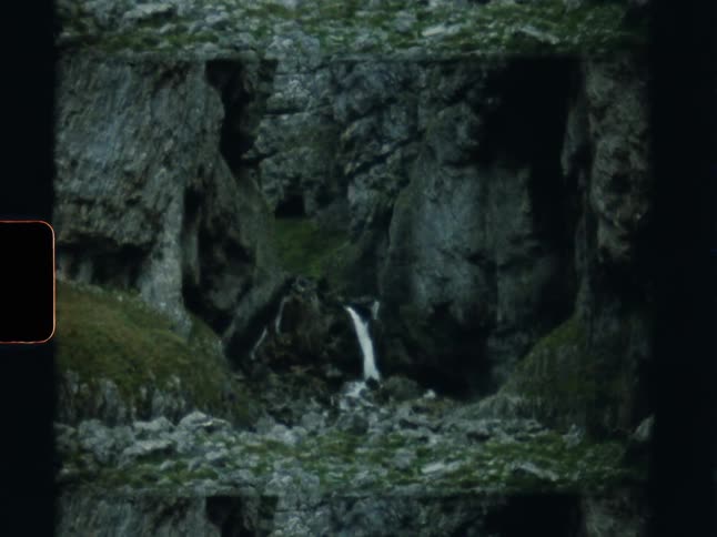 Archival 8mm footage shows a small waterfall cascading through a remote, rocky mountain gorge