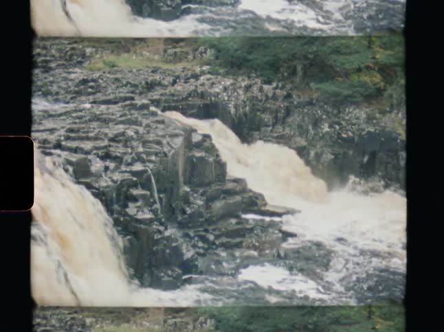 Vintage Super 8 footage shows a powerful waterfall rushing over dark, jagged rocks in a gorge