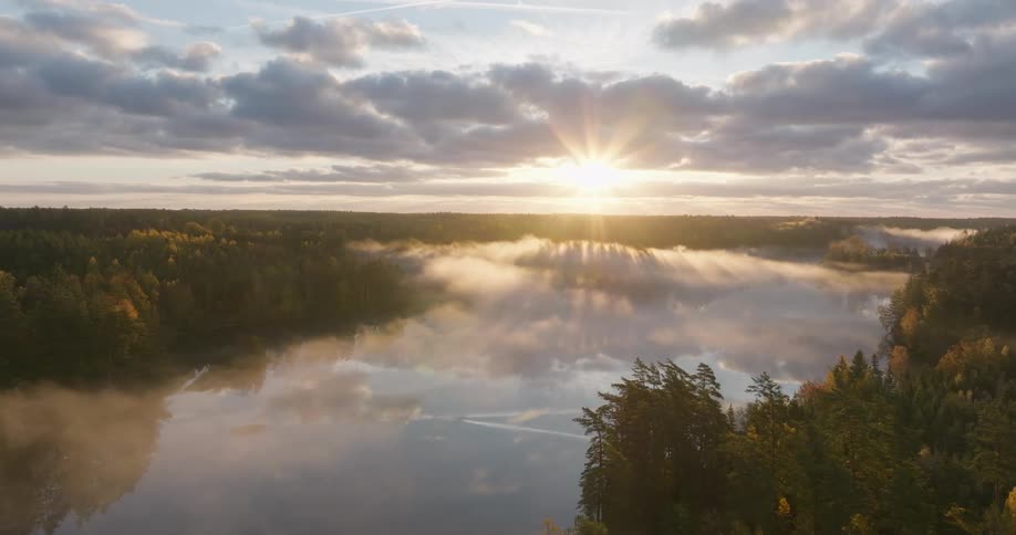 An epic aerial shot captures a beautiful sunrise over a misty lake and surrounding autumn forest, with sun rays breaking through the clouds