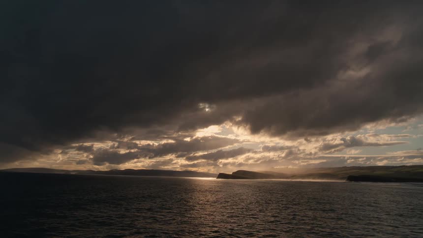 Dramatic storm clouds hang over the ocean as the setting sun breaks through, creating a golden reflection on the water