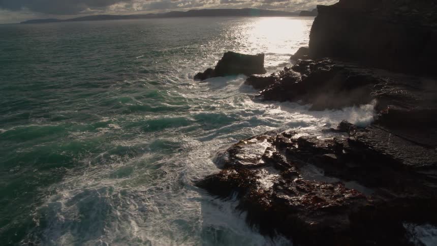 A dramatic slow-motion aerial shot captures turbulent ocean waves crashing against a rugged, rocky cliffside as the sun reflects on the water