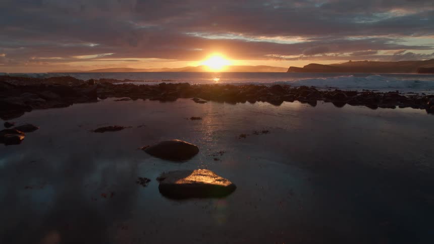 Dramatic aerial shot pulls back over a rocky coastline and tide pools during a stunning ocean sunset