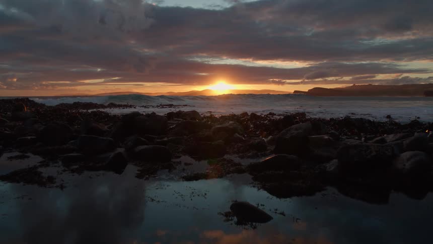 Slow motion tracking shot over tide pools and a rocky shoreline as waves crash during a dramatic sunset