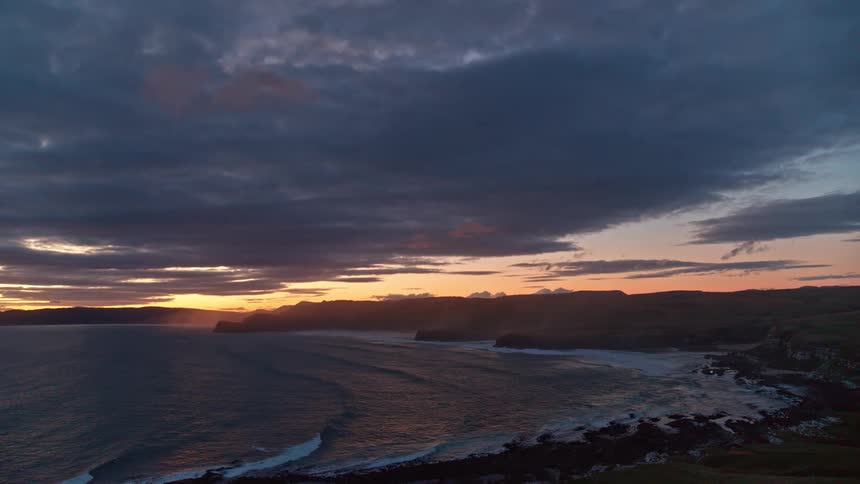A dramatic sunset casts a golden glow over a rugged coastline as waves roll into the shore under a cloudy sky