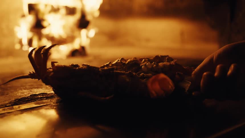 A chef places a fresh lobster dish into a hot wood-fired oven to cook next to the roaring flames