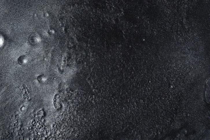 A close-up, abstract shot of a viscous black and silver metallic liquid as it slowly dries and solidifies, creating a granular texture