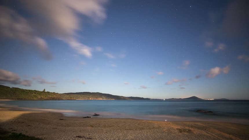 A stunning night time-lapse captures clouds and stars moving across the sky above a tranquil beach cove