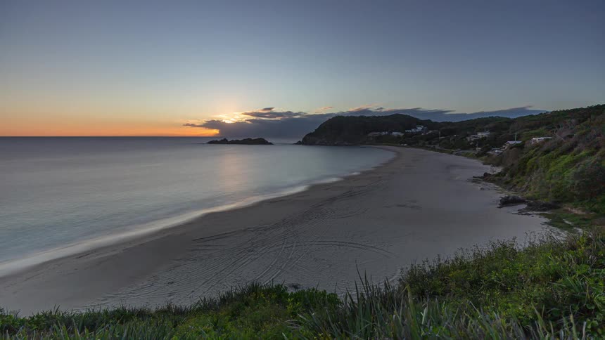 A serene timelapse captures the sun rising over a tranquil coastal bay, gradually illuminating the calm ocean and sandy shore