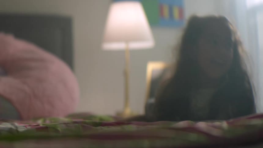 A smiling young girl with pigtails plays on her bed, with the camera focus shifting to a framed photo of her father on the nightstand
