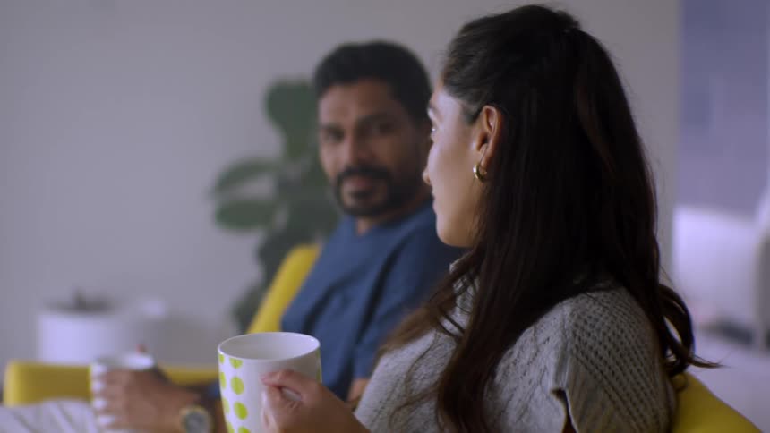A young woman listens to her partner while they enjoy a warm drink together on the sofa in their living room