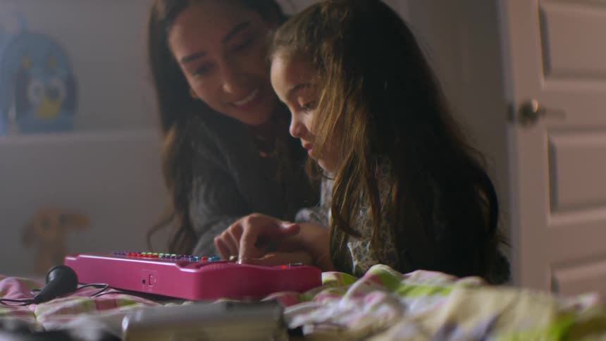 A loving mother smiles as she teaches her young daughter how to play a toy keyboard in her softly lit bedroom