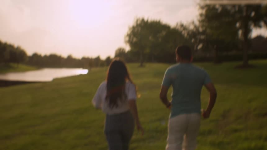 A happy family with a young daughter runs playfully together across a grassy field towards a lake at sunset