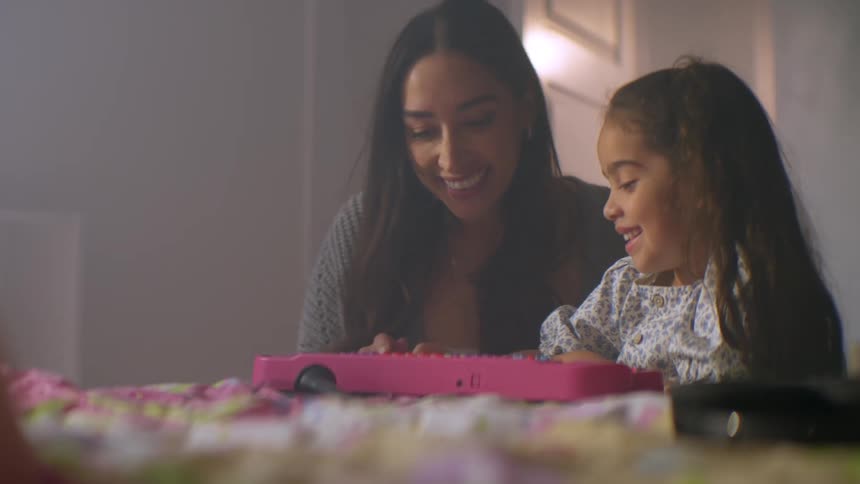 A loving mother joins her young daughter for a tender moment of playing together on a toy keyboard in a softly lit bedroom