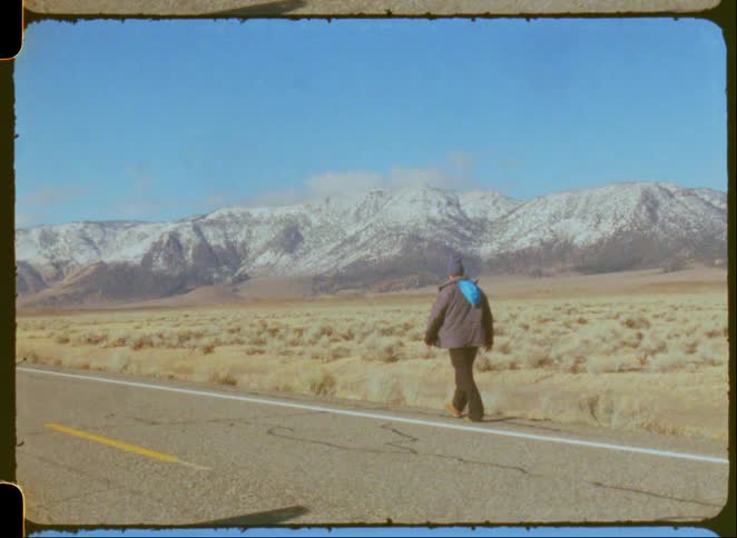 Vintage 8mm film captures a lone hiker walking down a deserted road towards snowy mountains on a clear day