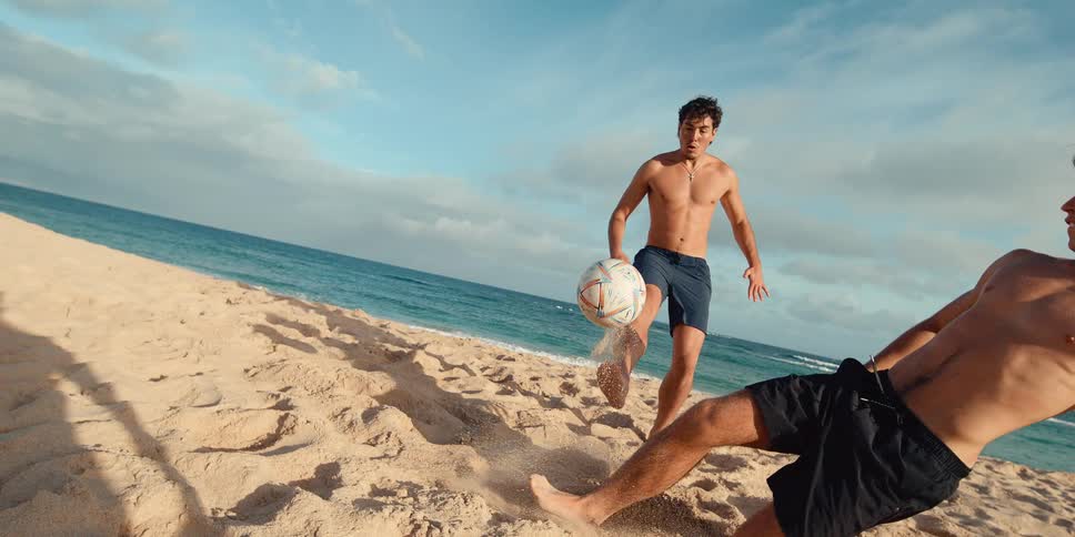 Two athletic men play an intense game of soccer on a sandy beach in dramatic slow motion