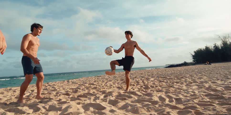 Friends have fun playing soccer on a sandy beach as one man skillfully juggles the ball in super slow motion