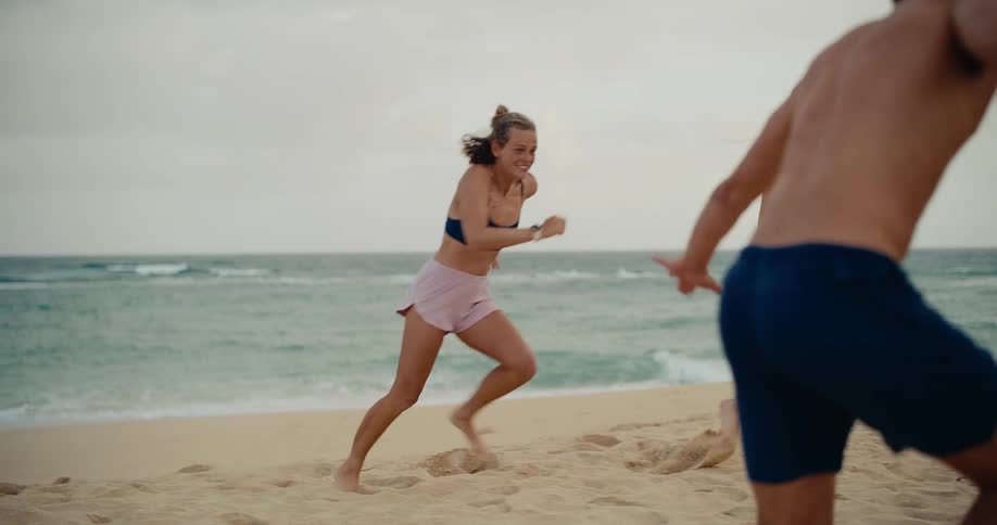 Three athletic young friends in swimwear laugh and run together on the beach in slow motion