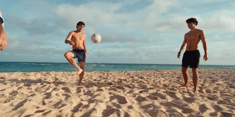 Two athletic young men play soccer on a sandy beach, with one juggling the ball on his foot in super slow motion