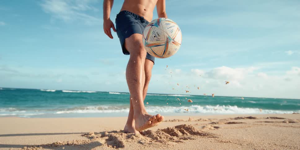 Close-up slow motion of a man balancing a soccer ball on his sandy foot on a tropical beach next to the ocean waves