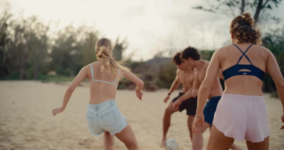 A group of active young friends play a competitive game of soccer on a sandy beach in slow motion