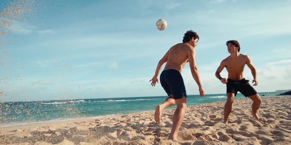 A man powerfully kicks a soccer ball on a sandy beach, sending a dramatic spray of sand into the air in stunning super slow motion