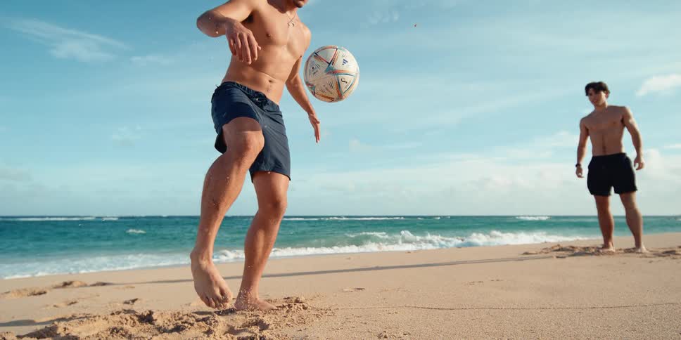 A man skillfully juggles a soccer ball on his foot in super slow motion while playing with a friend on a sunny beach