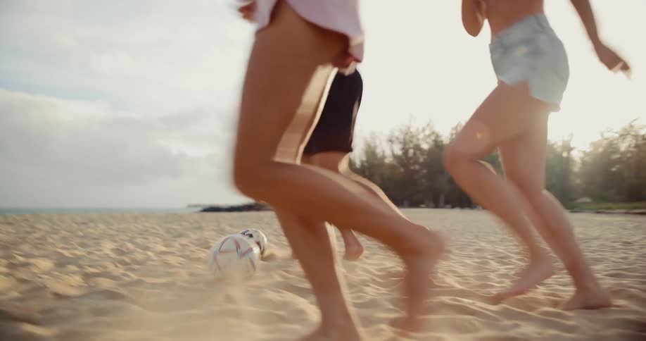 Energetic friends laugh and run while playing a game of soccer on the beach at sunset in slow motion