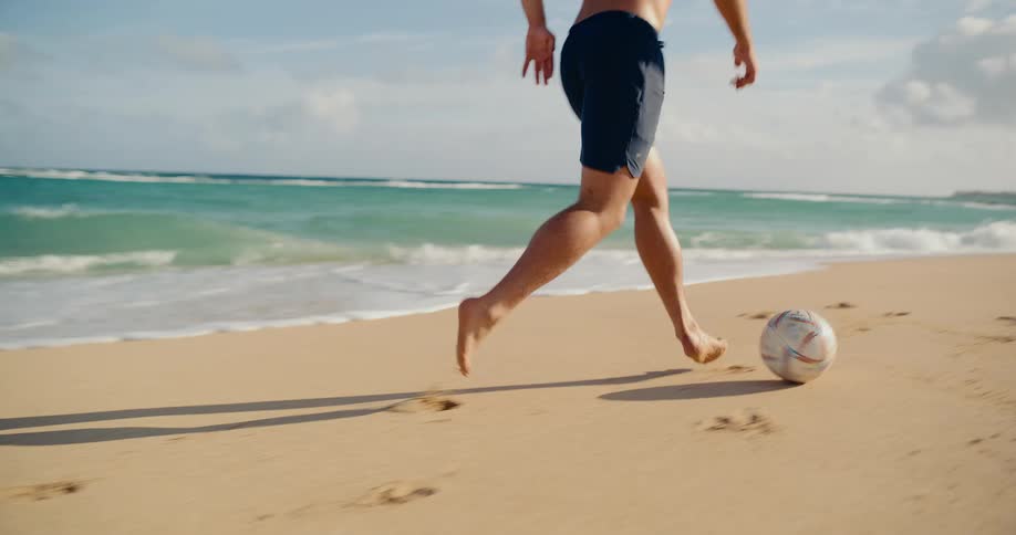 Athletic man plays soccer barefoot on a beautiful sandy beach, dribbling the ball along the shoreline in slow motion
