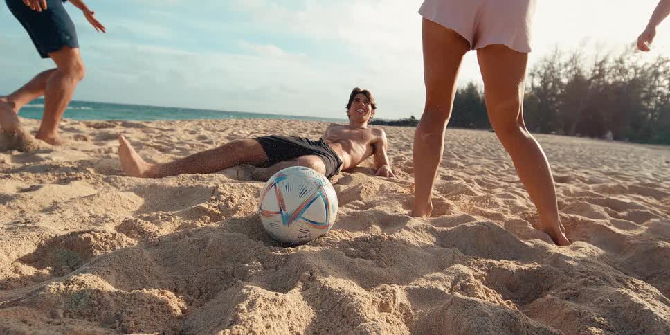 A soccer ball rests on the sand as grains fly through the air in slow motion while friends play on a sunny beach