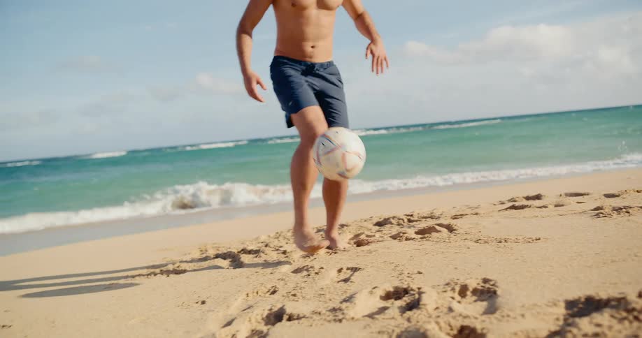 Slow motion shot of a fit young man juggling a soccer ball with his feet on the beach next to the ocean