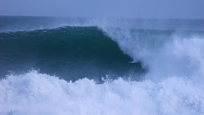 An expert surfer disappears into the barrel of a giant, powerful ocean wave in dramatic slow motion on a stormy day