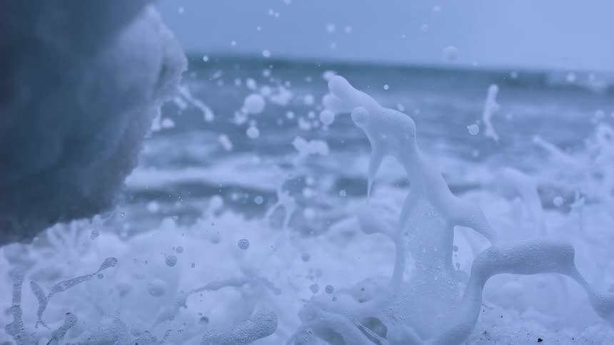 A dramatic super slow-motion close-up of a foamy ocean wave splashing against a snow-covered shore on a cold day