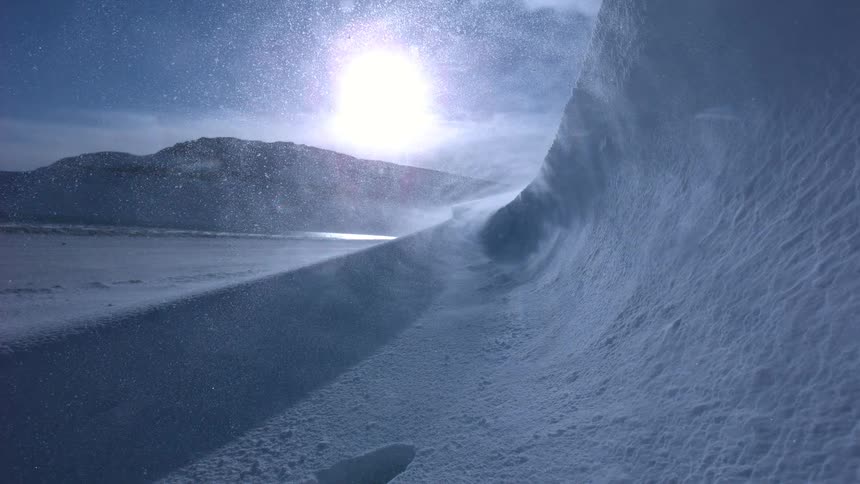 A dramatic slow-motion shot captures a fierce blizzard as wind whips snow across a vast, frozen landscape under a bright sun