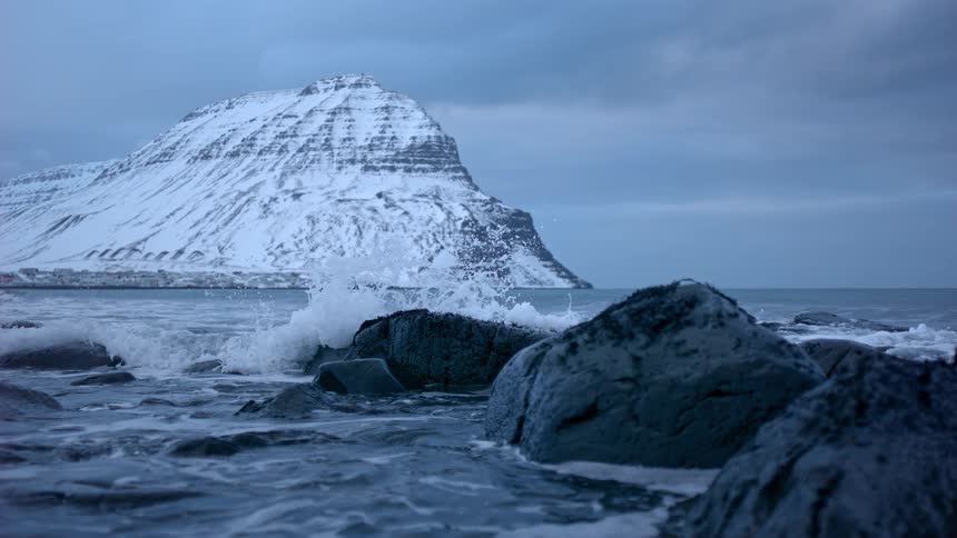 A cinemagraph captures a dramatic ocean wave frozen mid-splash against rocks with a majestic snow-covered mountain in the background