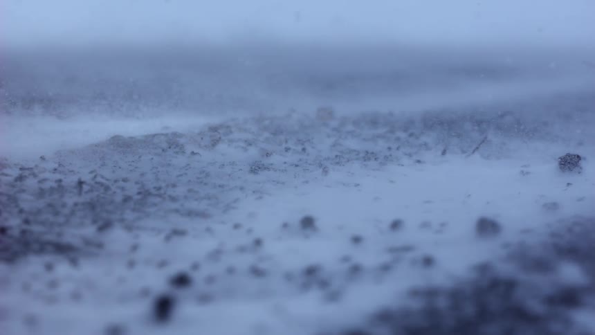 A low-angle close-up shot captures snow blowing across the frozen ground during a heavy winter blizzard