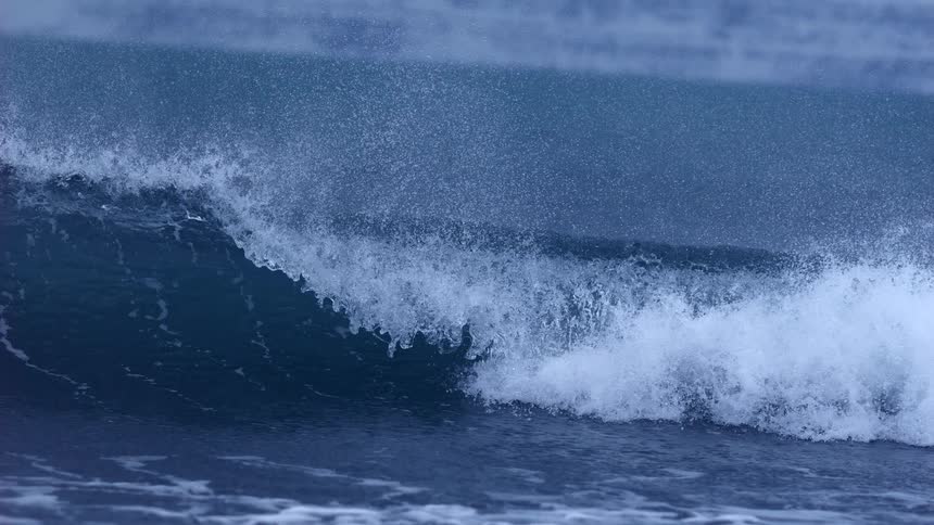 A dark blue ocean wave crests with white foam and spray in a dramatic super slow-motion close-up