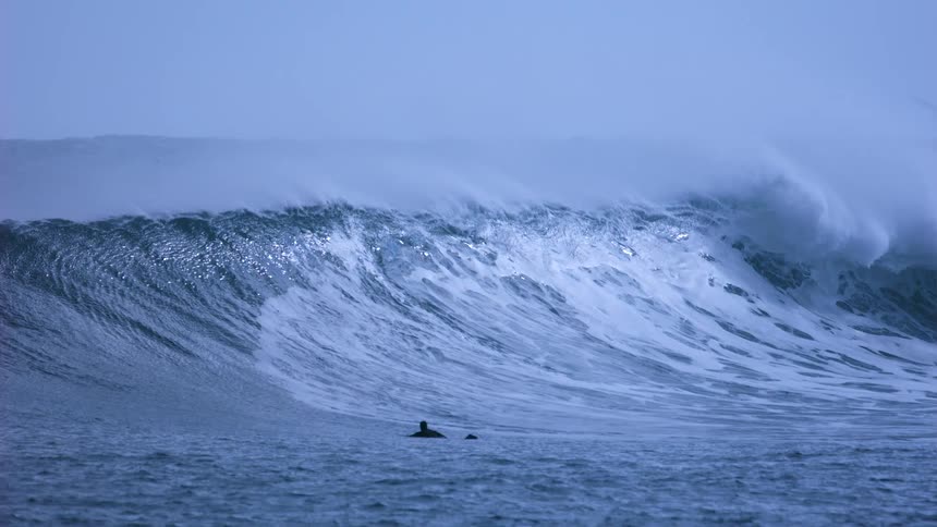 A lone surfer waits in the ocean as a massive, powerful wave crests in dramatic slow motion
