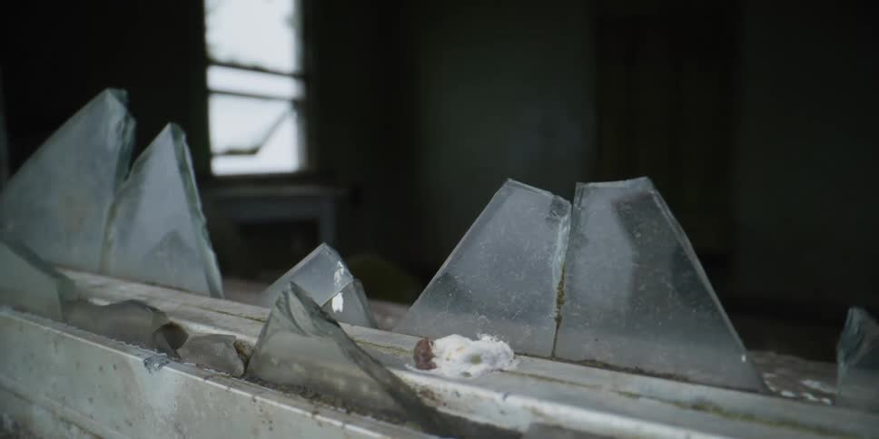 A slow pan reveals jagged shards of broken glass on a dirty windowsill in a dark, abandoned building