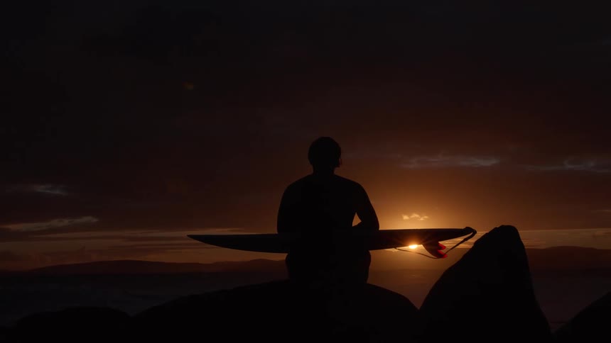 A silhouetted surfer sits on the rocks with a surfboard, contemplating the ocean as the sun rises on the horizon