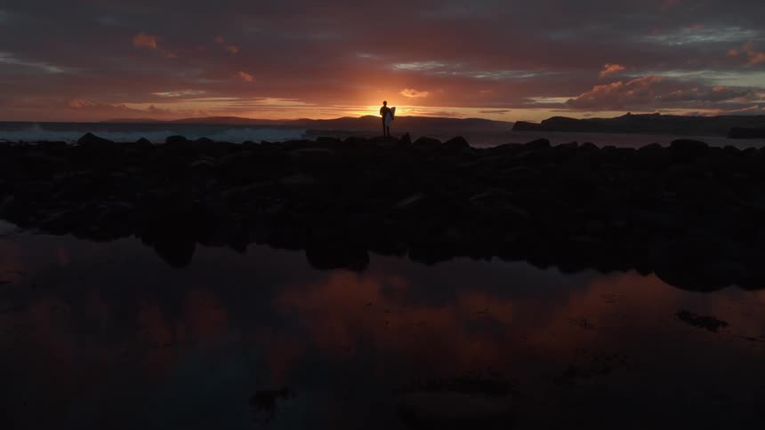 A lone surfer stands silhouetted on a rocky coastline, holding their surfboard while watching the vibrant sunset reflect in a still tide pool